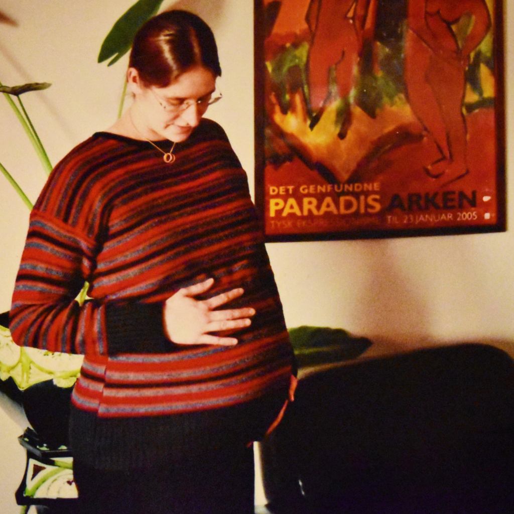 Image of a pregnant woman in a stripy jumper and black trousers, with her hands on her belly. I the background there's an art poster and a pot plant.